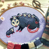 Front view of possum mama cross stitch pattern. The opossum mother has 6 babies, 5 of which are on her back. The 6th one is sitting on her left. Colors are mainly blueish grey, lilac, beige and pink. The baby possums are extremely cute.