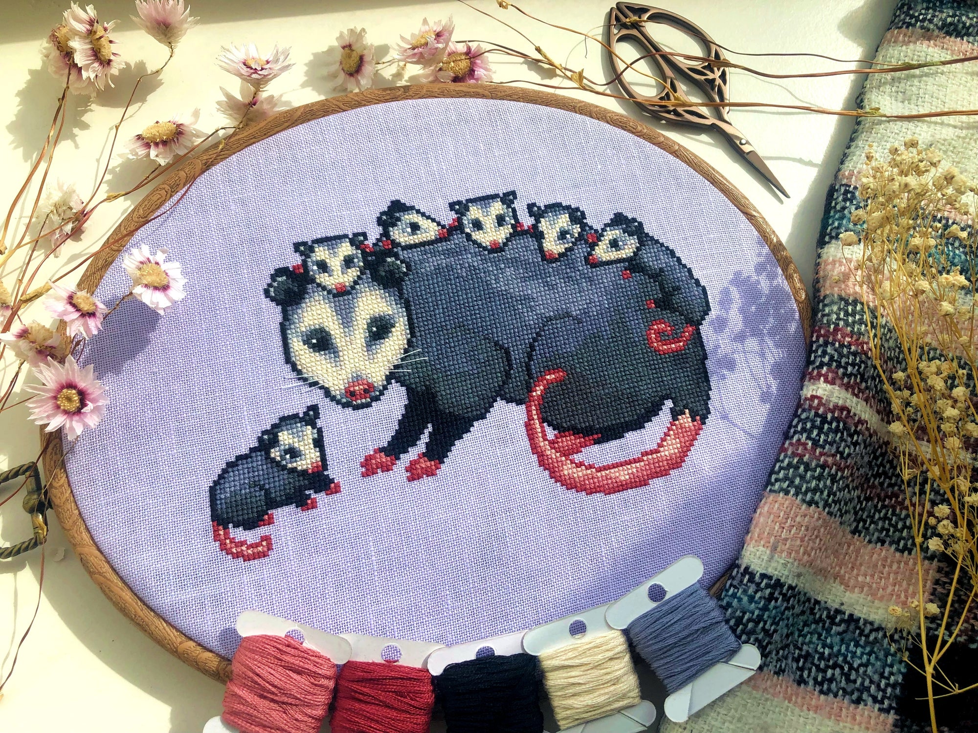 Front view of possum mama cross stitch pattern. The opossum mother has 6 babies, 5 of which are on her back. The 6th one is sitting on her left. Colors are mainly blueish grey, lilac, beige and pink. The baby possums are extremely cute.