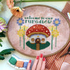 Front view of Fungalow mushroom house cross stitch pattern. Green quote reads Welcome to Our Fungalow, that is placed above the scene. Piece is framed in oval embroidery hoop, and is surrounded by decorations. Colors are red, green, yellow, and lilac