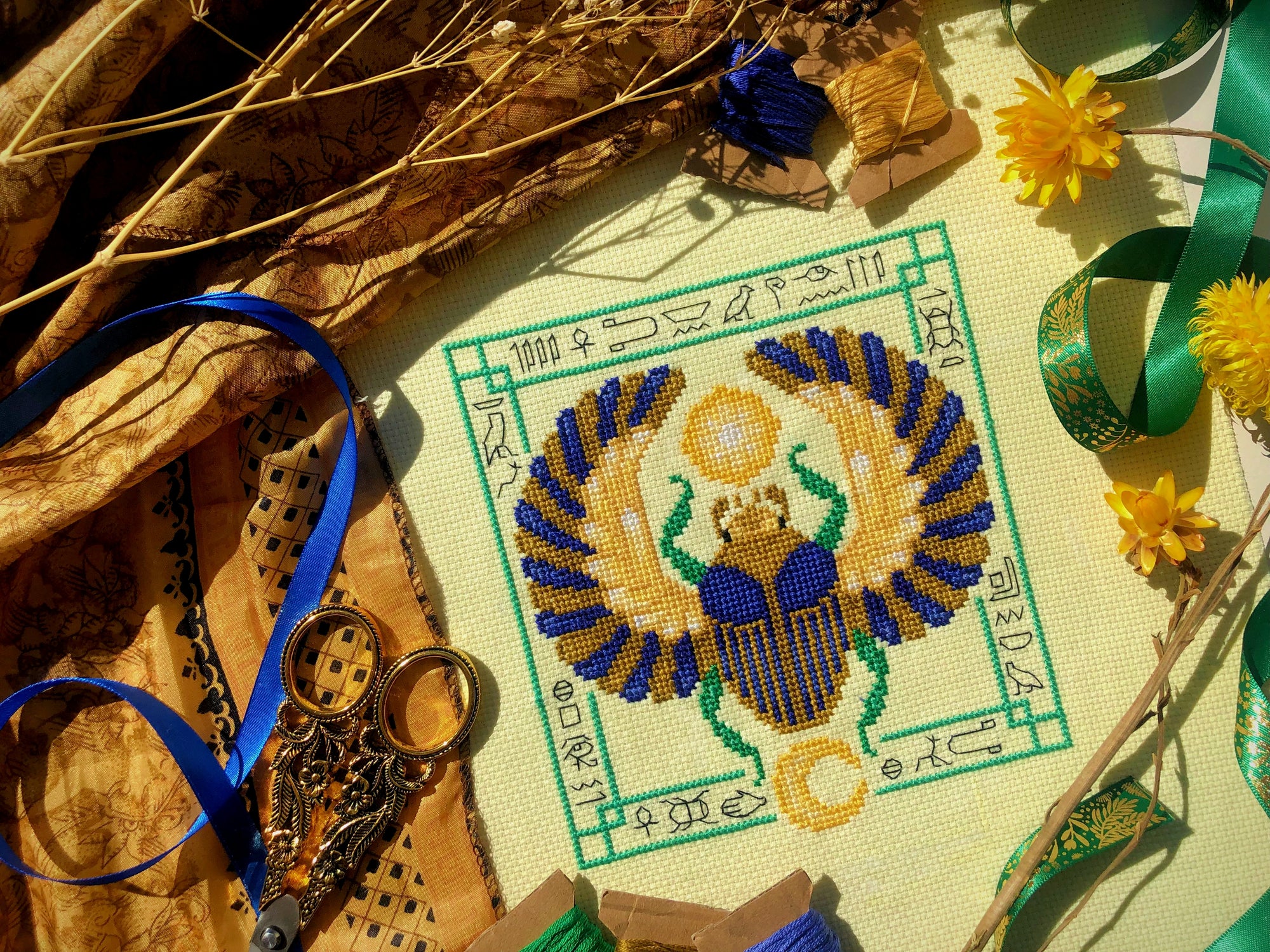 Top down, angled view of Egyptian Scarab cross stitch pattern by NeedleLot Designs. Colors are blue, old gold, green, black and yellow. Piece consists only of DMC threads. Scarab is flanked by sun and moon, and is surrounded by hieroglyphs.