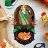 Vertical flat-lay of Sleeping Fox cross stitch pattern. Stitched item, surrounded by decorations. Finished piece is of medium size. Colors are green, brown, orange, black, beige and white. Fox is sleeping underground. Trees and sun rays are above.