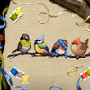 Flatlay of Winter Birds cross stitch pattern. Stitched item is surrounded by decorations. Finished piece is of medium to large size. Colors are yellow, brown, orange, blue, green, white. There are four birds sitting on a branch wearing winter gear.