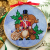 Front view of Nutcracker squirrel cross stitch pattern. Squirrel is wearing a military suit and looks stern. His jacket is red and he is wearing black boots and a hat. Around him are walnuts, acorns, holly and a bauble. Scene is surrounded by decor.