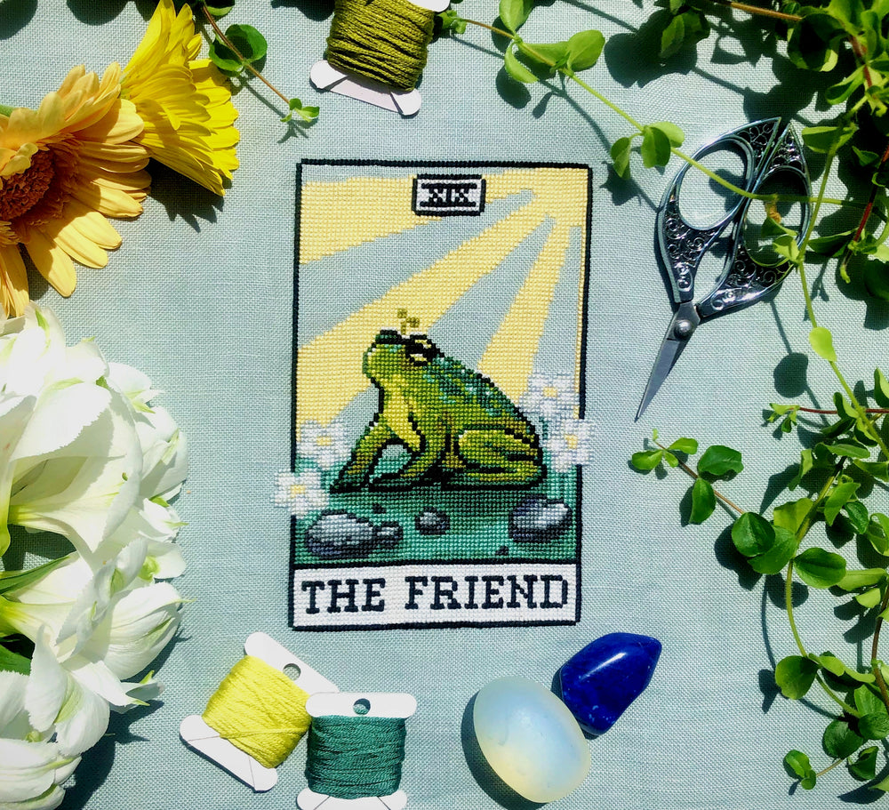 Flatlay of The Friend frog tarot cross stitch pattern. Stitched item, surrounded by decorations. Finished piece is of small to medium size. Colors are yellow, green, blue, grey and black. Frog is cute and small. The Friend is written underneath.