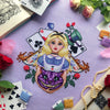 Alice in Wonderland cross stitch pattern front view. Piece is large and detailed. Alice is in the middle of the composition, holding the head of the Cheshire Cat in her arms. Alices hair is blonde with a black ribbon, her dress is blue and white.
