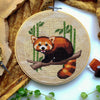 Flat-lay of Red Panda cross stitch pattern. Stitched item, surrounded by foliage, scissors and decor. Finished piece is of small to medium size. Colors are yellow, brown, green and white. Red Panda is cute and small and is looking at the viewer.