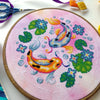 Flat-lay of Koi Fish cross stitch pattern. Stitched item, surrounded by decorations. Finished piece is of medium size. Colors are very bright, and are green, blue, orange, pink and yellow. There are two fish, surrounded by lotus leaves and flowers.