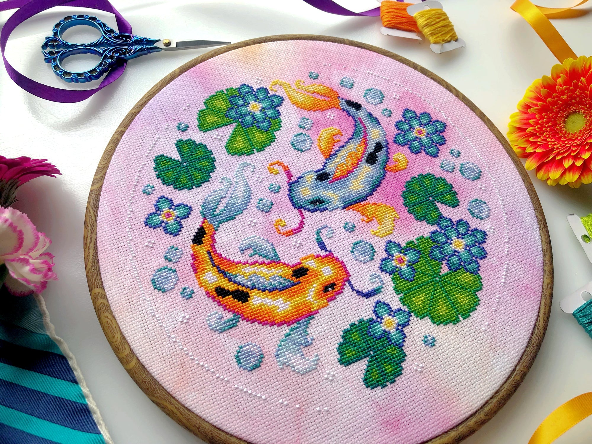 Flat-lay of Koi Fish cross stitch pattern. Stitched item, surrounded by decorations. Finished piece is of medium size. Colors are very bright, and are green, blue, orange, pink and yellow. There are two fish, surrounded by lotus leaves and flowers.