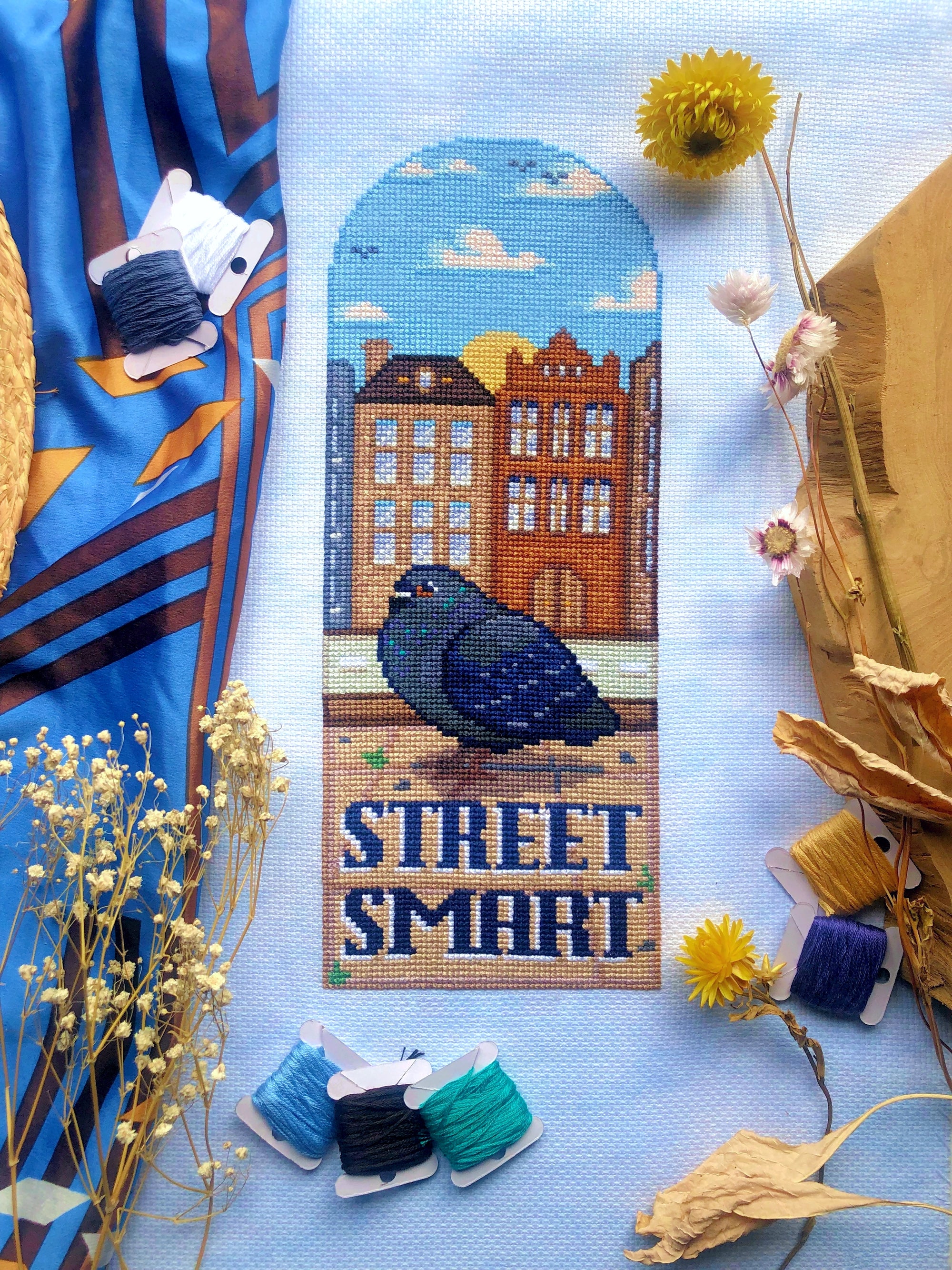 Full front view of Street Smart Pigeon cross stitch pattern. Scene is surrounded by decorations. Pigeon is rather fat, and is standing on a sidewalk. Behind him are Amsterdam houses. Underneath the pigeon are the words Street Smart. Scene is colorful