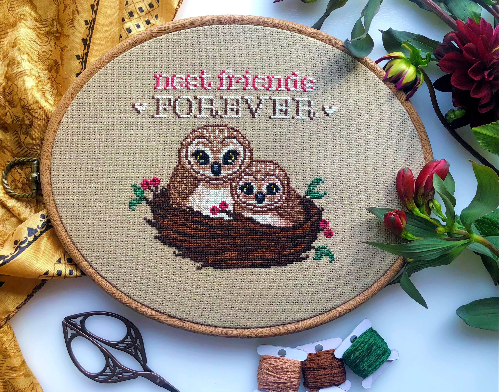 Front view of Nest Friends Forever owls cross stitch pattern. Two owls are nestled together in a nest made of branches and berries. There is one small owl and one big owl. Above them is the quote Nest Friends Forever. The birds are brown and cute.