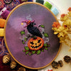 Flat-lay of Jack-Crow-Lantern cross stitch pattern. Stitched item, surrounded by decorations. Finished piece is small. Colors are orange, purple, black, and green. Crow is wearing a hat and looking over its shoulder, standing on a smiling pumpkin.