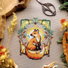 Flat-lay of Art Nouveau Fox cross stitch pattern. Stitched item, surrounded by decorations. Finished piece is of small to medium size. Colors are yellow, brown, orange, green and white. Fox is cute and small and is looking over its shoulder.