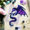 Flat-lay of Dragon cross stitch pattern. Stitched item, surrounded by decorations. Finished piece is of medium to large size. Colors are blue, purple, white, black and pink. Dragon is surrounded by variegated frame. Its head is like that of a horse.