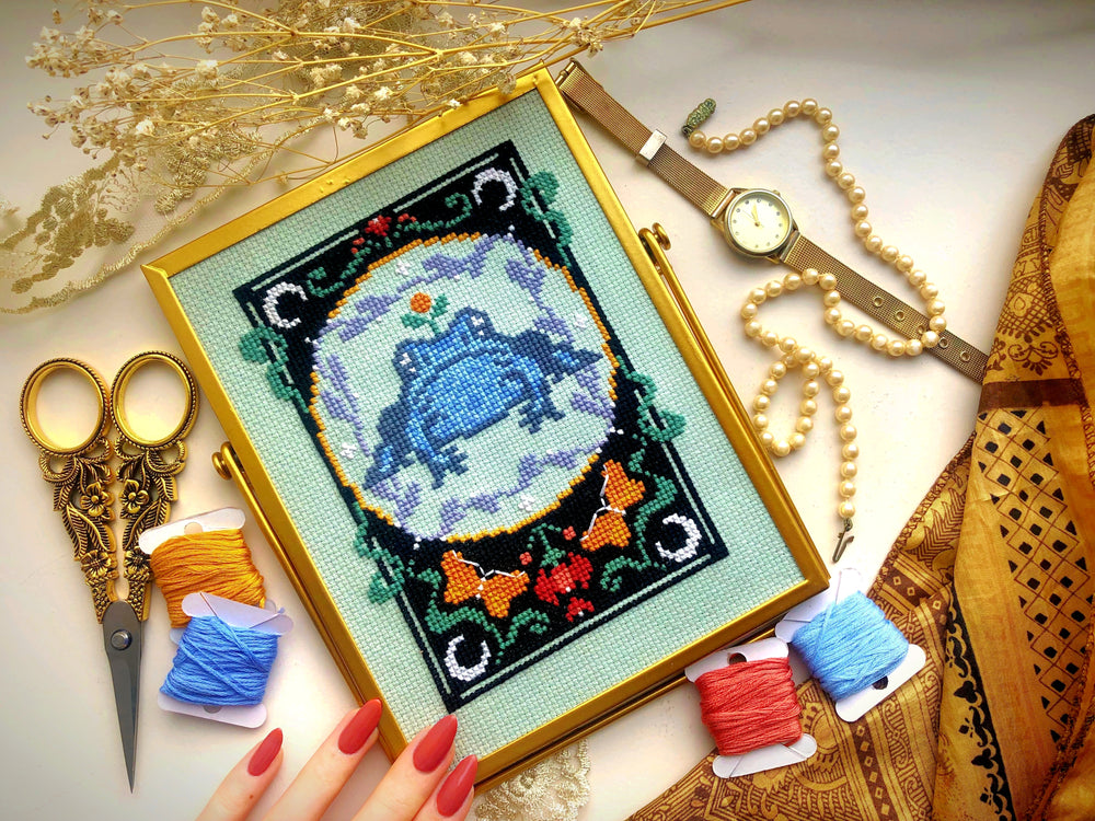 Front view of Art Nouveau frog cross stitch pattern. Scene is surrounded by golden decorations. Frog is blue and grey, and is placed in a black frame with green, red, purple and orange floral decorations. There are also moons and butterflies.