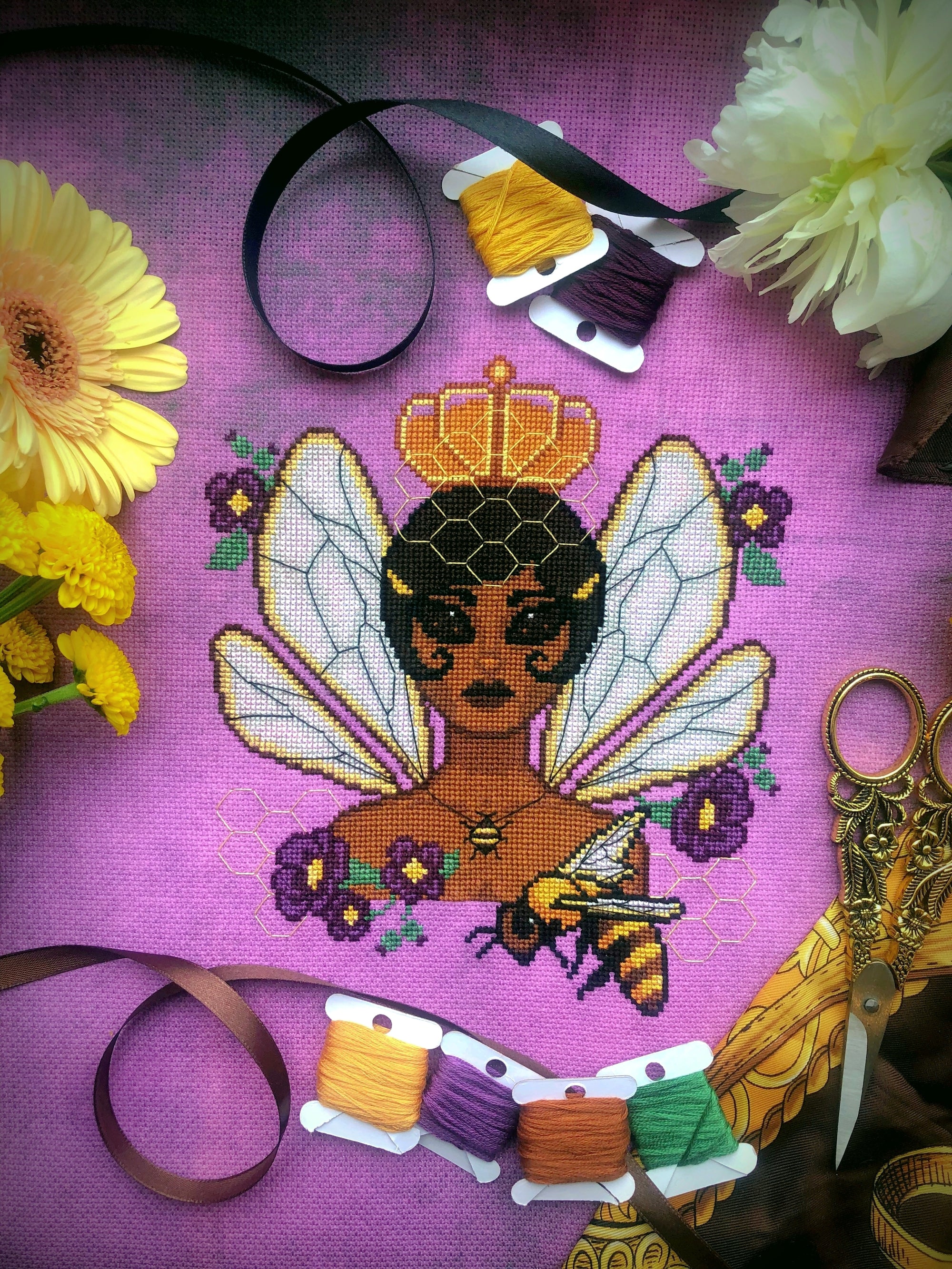 Vertical front view of Queen Bee cross stitch pattern. Queen Bee is represented as a black woman with dark hair that is cut into a bob. She is wearing an orange crown and has the wings of a bee. She wears a necklace and is surrounded by flowers.