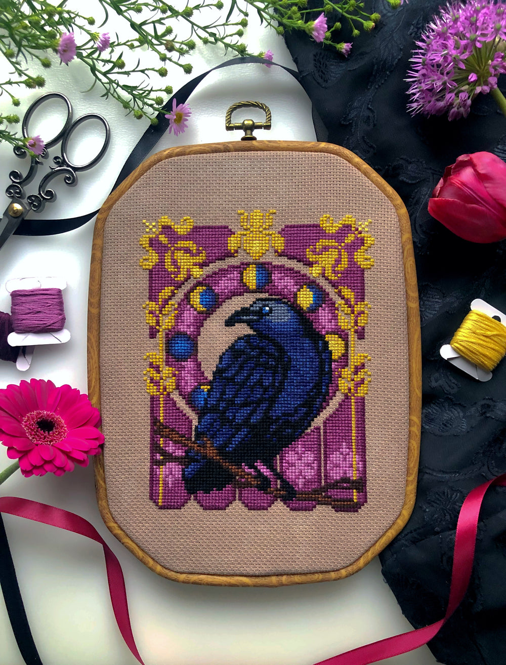 Flat-lay of Art Nouveau Crow cross stitch pattern. Stitched item in hoop, surrounded by decorations. Finished piece is of small to medium size. Colors are yellow, brown, black, blue and purple. Crow is looking over its shoulder.