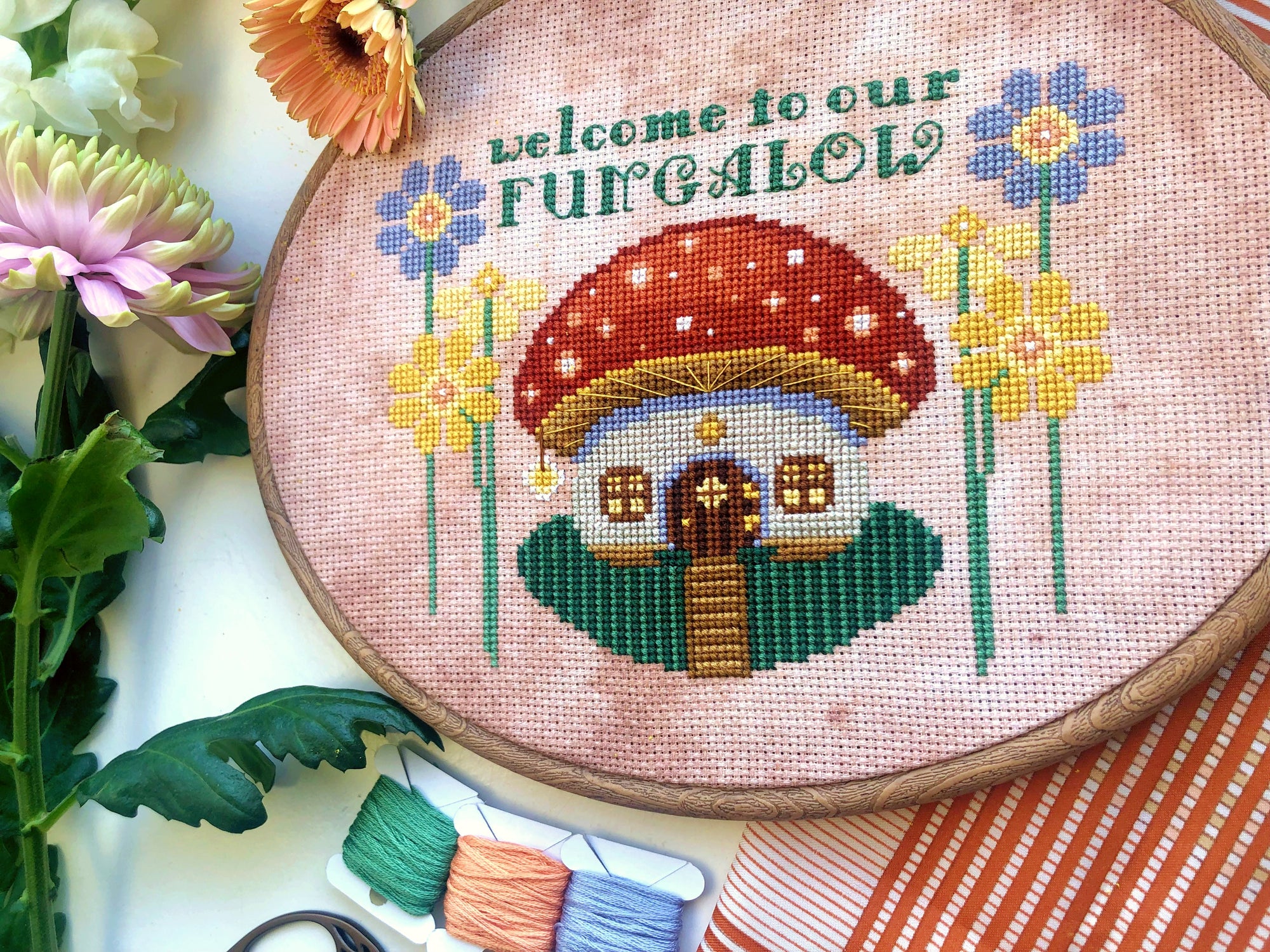 Closer view of toadstool mushroom house cross stitch pattern. House is framed on either side by yellow and lilac flowers. The house has a little round door and several windows. In front is a path, surrounded by green grass. There is a lantern.