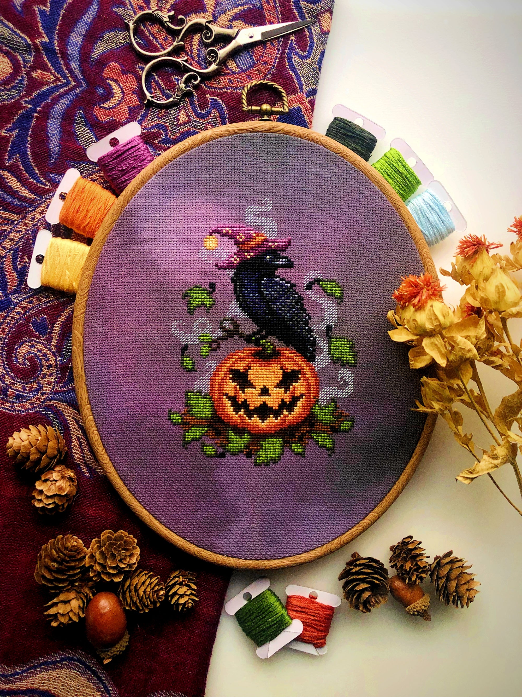 Flat-lay of Jack-Crow-Lantern cross stitch pattern. Stitched item, surrounded by decorations. Finished piece is small. Colors are orange, purple, black, and green. Crow is wearing a hat and looking over its shoulder, standing on a smiling pumpkin.