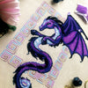 Flat-lay of Dragon cross stitch pattern. Stitched item, surrounded by decorations. Finished piece is of medium to large size. Colors are blue, purple, white, black and pink. Dragon is surrounded by variegated frame. Its head is like that of a horse.