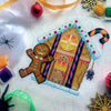 Flat-lay of Cookies & Scream gingerbread house cross stitch pattern, at a slight angle. Finished piece is of medium size. Piece is bright and colorful, eminating both Christmas spirit and Halloween spirit. Candy and candycanes are everywhere.