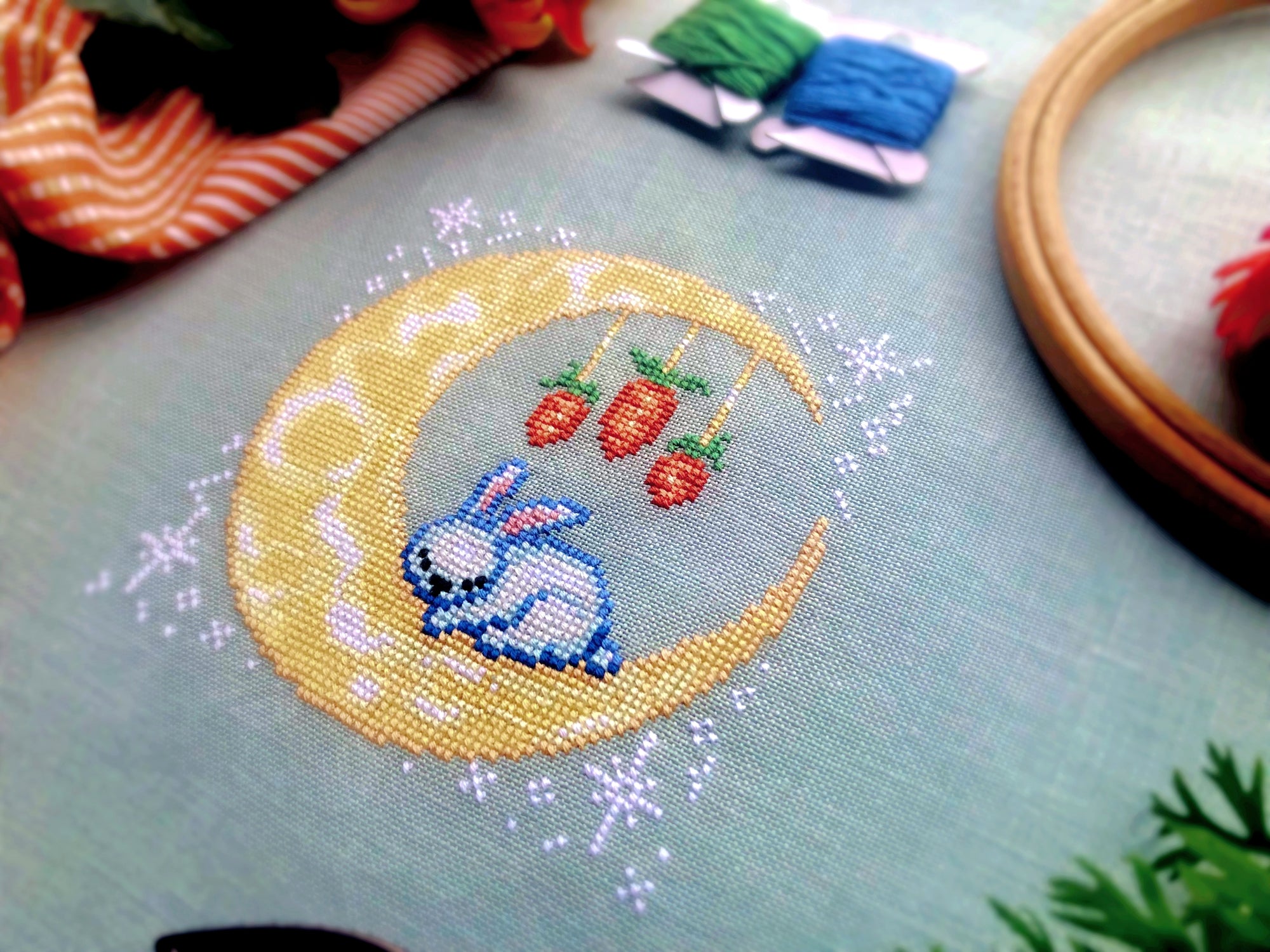 Closeup of sleeping bunny cross stitch pattern, laying on a yellow moon. Bunny is white with blue and pink details. He is very small. The moon is yellow with color variations. From the moon are carrots hanging down, like a baby mobile.