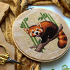 Flat-lay of Red Panda cross stitch pattern. Stitched item, surrounded by foliage, scissors and decor. Finished piece is of small to medium size. Colors are yellow, brown, green and white. Red Panda is cute and small and is looking at the viewer.