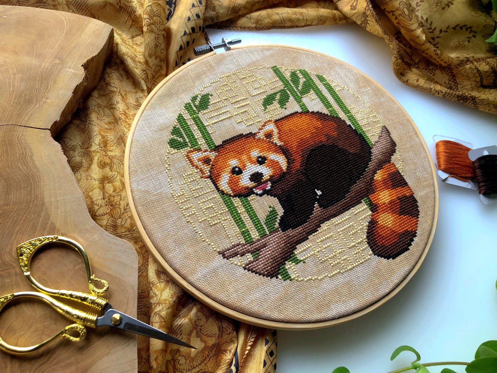 Flat-lay of Red Panda cross stitch pattern. Stitched item, surrounded by foliage, scissors and decor. Finished piece is of small to medium size. Colors are yellow, brown, green and white. Red Panda is cute and small and is looking at the viewer.