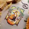 Flat-lay of Art Nouveau Fox cross stitch pattern. Stitched item, surrounded by decorations. Finished piece is of small to medium size. Colors are yellow, brown, orange, green and white. Fox is cute and small and is looking over its shoulder.