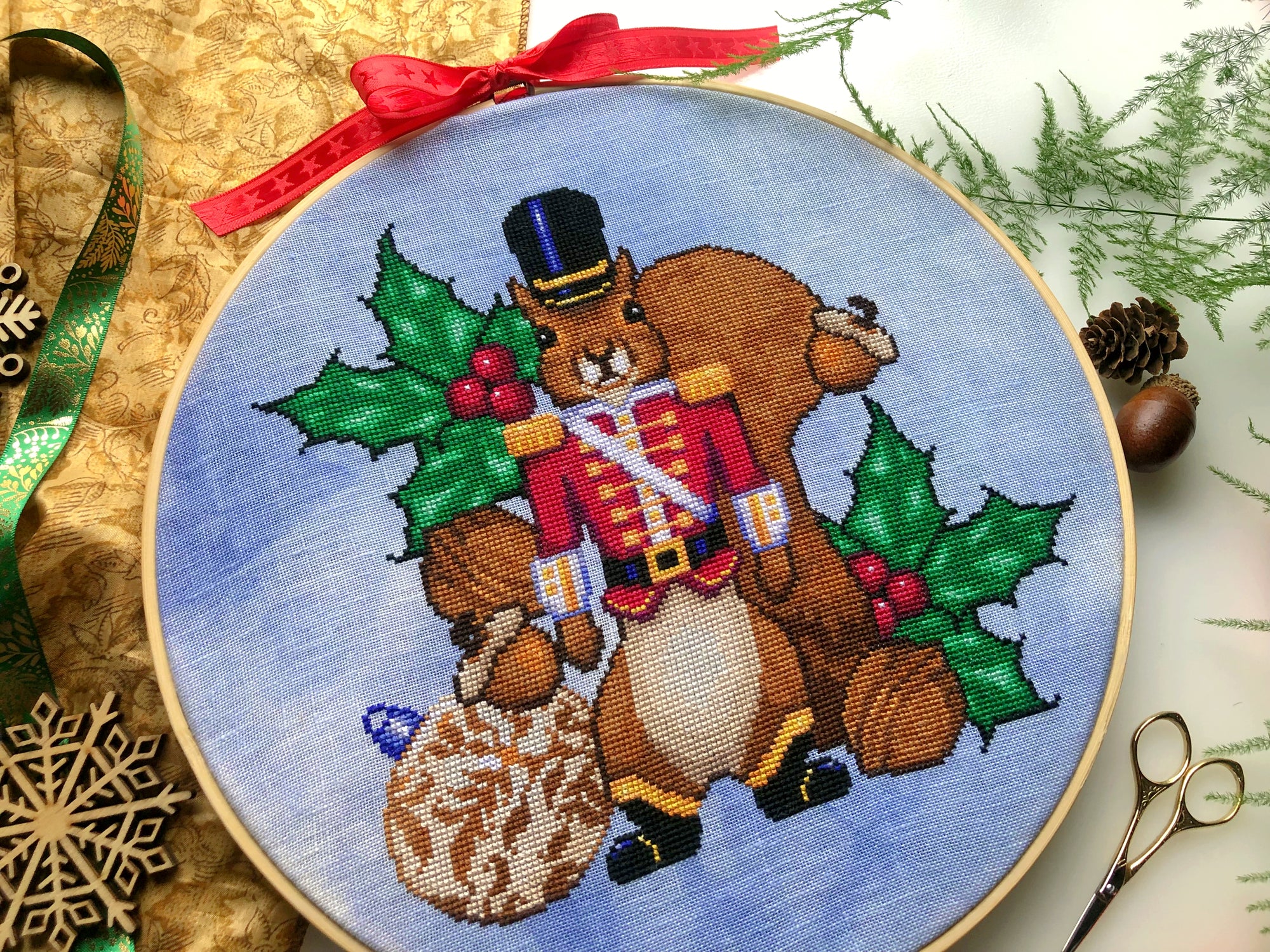 Angled front view of squirrel Nutracker cross stitch pattern. Colors are primarily blue, green, brown, red, purple, yellow and black. There are lots of details in all the elements. Squirrel looks realistic. He has a big tail and wears no pants.