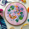 Flat-lay of Koi Fish cross stitch pattern. Stitched item, surrounded by decorations. Finished piece is of medium size. Colors are very bright, and are green, blue, orange, pink and yellow. There are two fish, surrounded by lotus leaves and flowers.