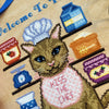 Closeup of cat baker. He is wearing a white chef's hat, and is wearing a checkered apron that says "Kiss the Chef". Colors are orange, brown, pink, purple, red, yellow and blue. Cat's eyes are blue, and he has whiskers. Stitches are neat and tidy.