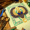 Top down, angled view of Egyptian Scarab cross stitch pattern by NeedleLot Designs. Colors are blue, old gold, green, black and yellow. Piece consists only of DMC threads. Scarab is flanked by sun and moon, and is surrounded by hieroglyphs.