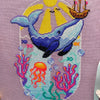 Font view of Whale with Boat cross stitch pattern. Blue whale has a boat on its nose that it has lifted out of the water. The sun is shining. Underwater we can see coral, fish, seaweed and a jellyfish. Colors are very vibrant. Birds are in the sky.