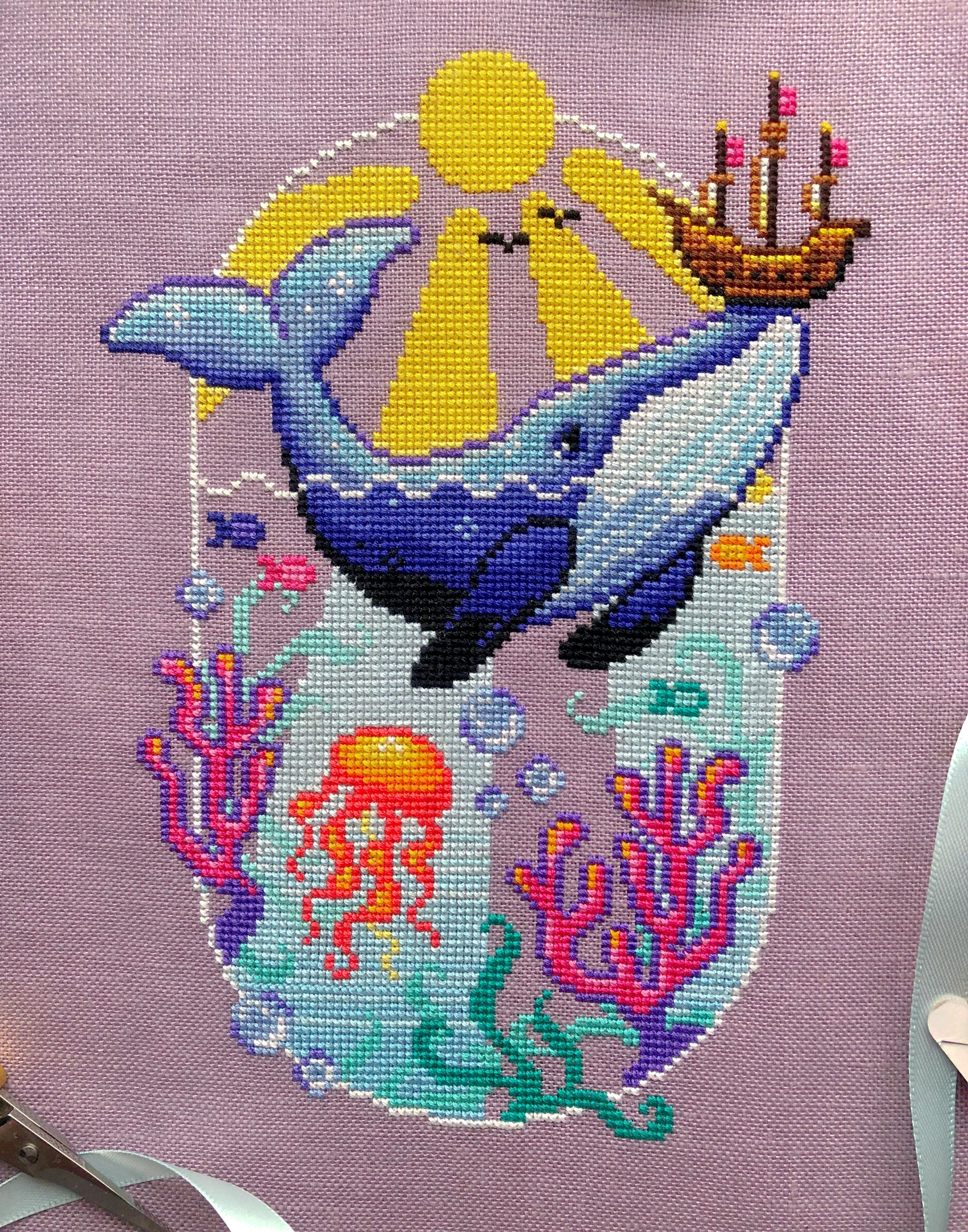 Font view of Whale with Boat cross stitch pattern. Blue whale has a boat on its nose that it has lifted out of the water. The sun is shining. Underwater we can see coral, fish, seaweed and a jellyfish. Colors are very vibrant. Birds are in the sky.