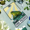 Flatlay of The Friend frog tarot cross stitch pattern. Stitched item, surrounded by decorations. Finished piece is of small to medium size. Colors are yellow, green, blue, grey and black. Frog is cute and small. The Friend is written underneath.