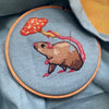 Image of the rat with mushroom umbrella being stitched. Stitches are neat and tidy. Fabric is placed inside an embroidery hoop. Colors are bright and beautiful: pink, brown, red, orange and yellow. Rat is looking cute and happy.