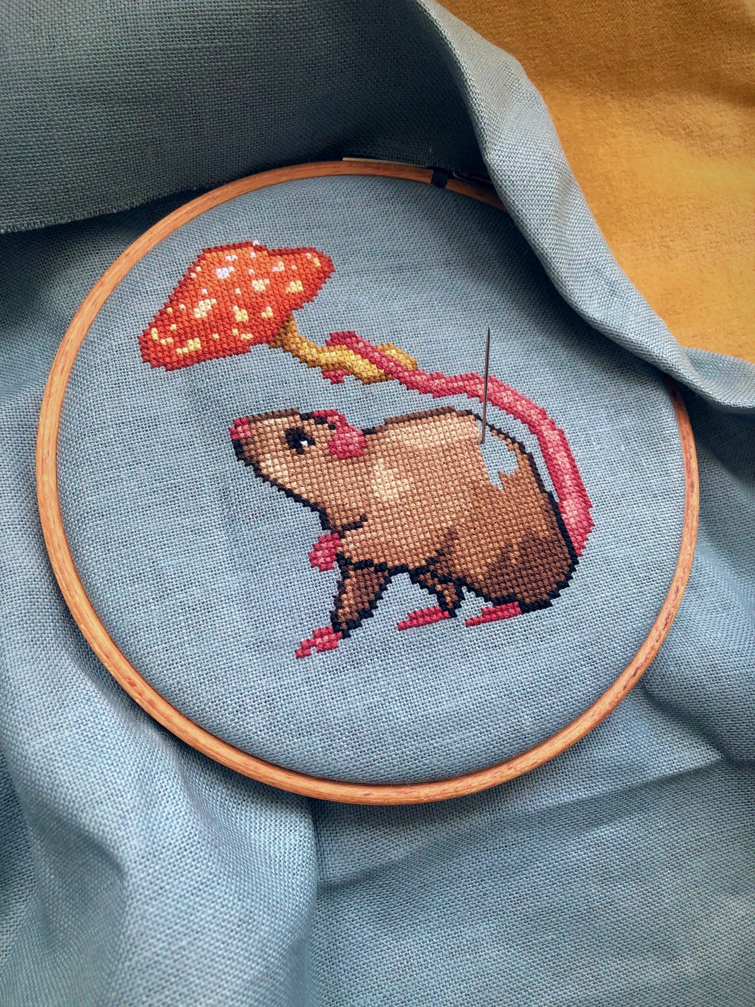 Image of the rat with mushroom umbrella being stitched. Stitches are neat and tidy. Fabric is placed inside an embroidery hoop. Colors are bright and beautiful: pink, brown, red, orange and yellow. Rat is looking cute and happy.