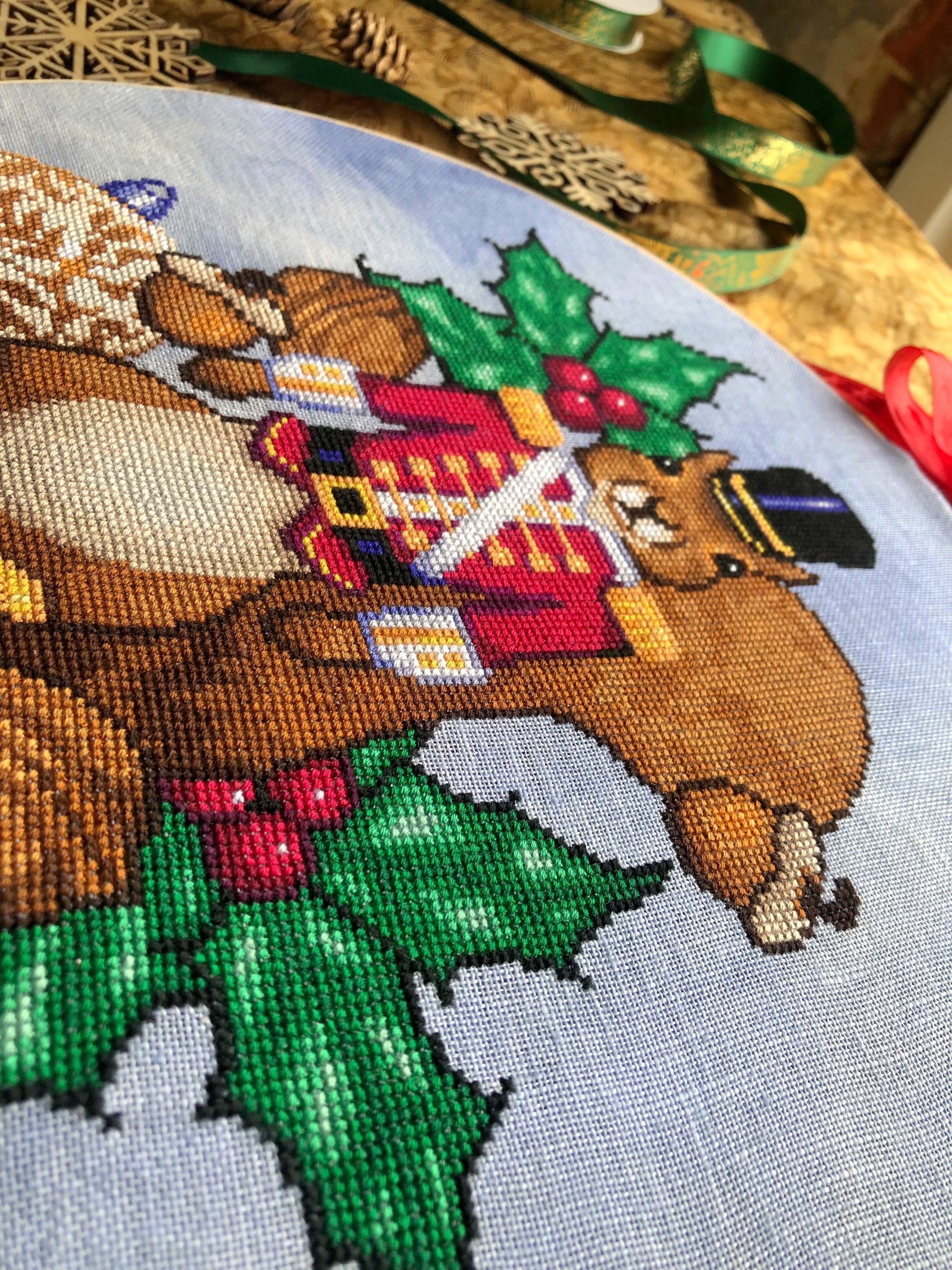 Closeup of squirrel cross stitch pattern, bottom righthand side. Stitches are neat and tidy. The pattern is rather large. Colors are vibrant and saturated. Especially the greens in the holly pop.