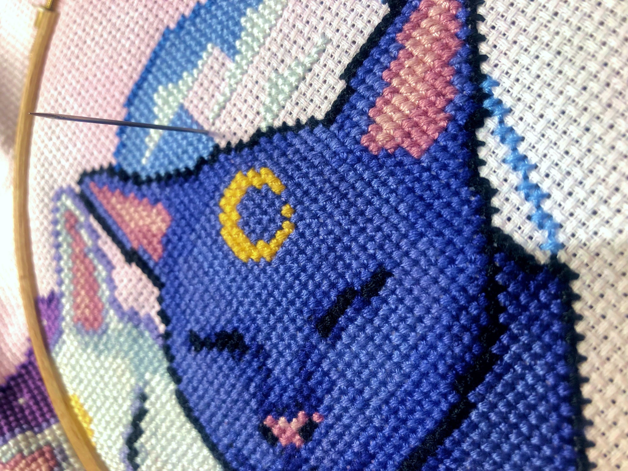 Extreme closeup of right-hand side of Smitten Kittens cross stitch pattern, and picture of the work in progress. Needle is sticking upwards. Danish method of stitching is being used. Moon on cats head is a vibrant yellow. Cat is cornflower blue.