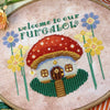 Closer view of toadstool mushroom house cross stitch pattern. House is framed on either side by yellow and lilac flowers. The house has a little round door and several windows. In front is a path, surrounded by green grass. There is a lantern.