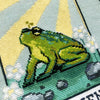 Closeup of The Friend frog tarot cross stitch pattern. Stitched item, surrounded by decorations. Finished piece is of small to medium size. Colors are yellow, green, blue, grey and black. Frog is cute and small. The Friend is written underneath.