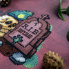 Closeup of R.I.B. cross stitch pattern, right bottom view. Stitches are neat and tidy. Frog hand is clearly visible. Tombstone is a brown/purple shade. It has a cross on top. Scene is surrounded by rocks and graveyard fence. Skull eyes are lit up.
