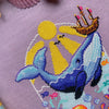 Closeup of Whale with Boat cross stitch pattern. Stitches are neat and tidy. There are little detailsin the wale, like shimmers. The boat is brown with sails and pink flags. The sun rays permeate the water. Bubbles are placed on the sides.