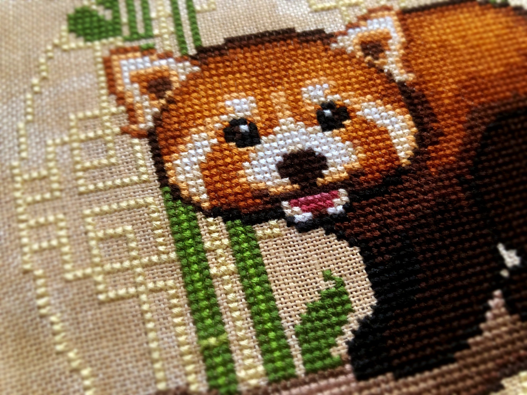 Closeup of Red Panda cross stitch pattern. The red pandas face is cute and round, and the scene is brightly colored. The details of the Chinese-inspired background are clearly visible. Bamboo shoots are placed behind the red pandas head and ears.