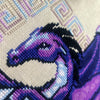 Closeup of Dragon cross stitch pattern. Stitched item, surrounded by decorations. Finished piece is of medium to large size. Colors are blue, purple, white, black and pink. Dragon is surrounded by variegated frame. Its head is like that of a horse.