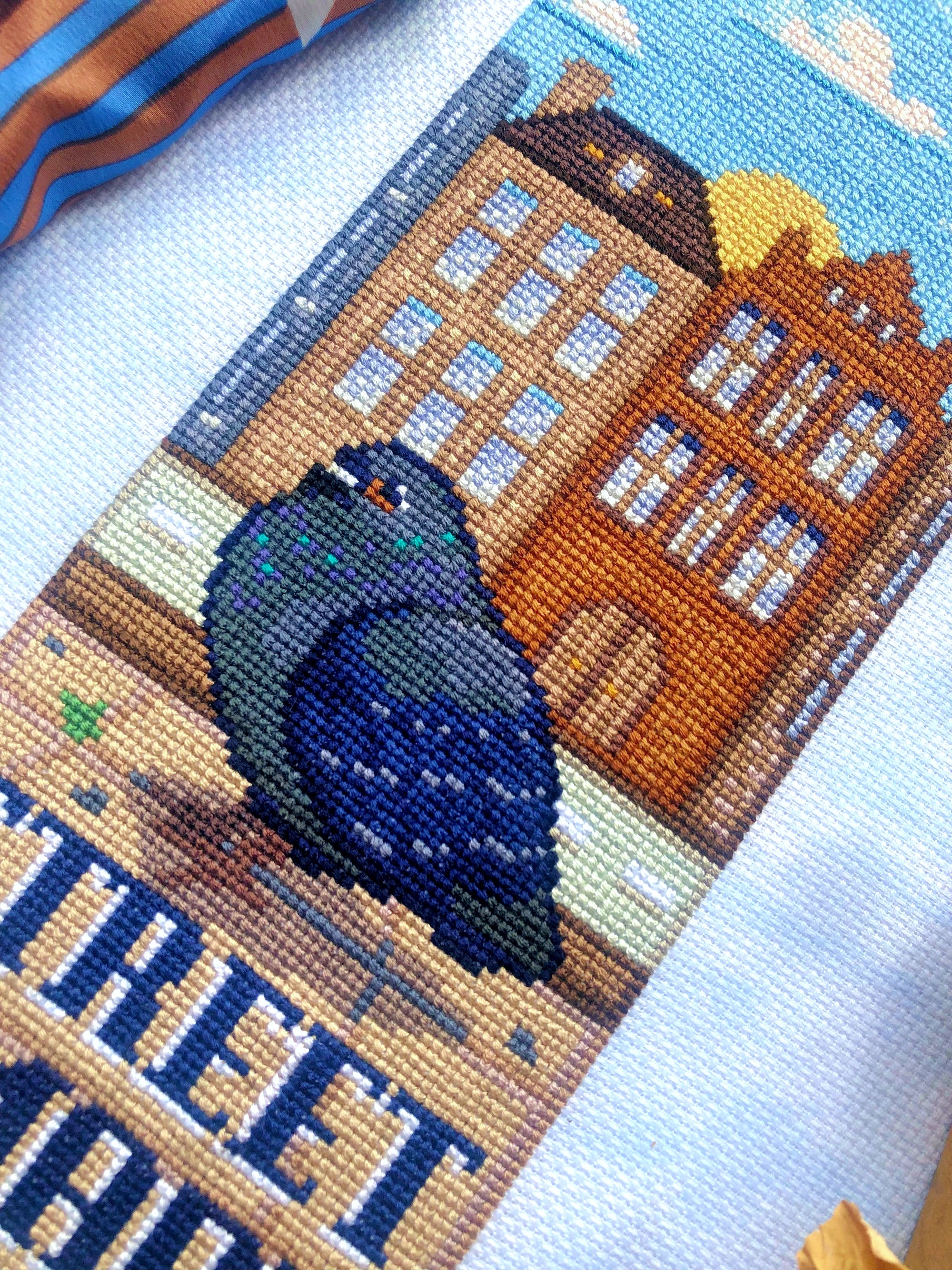 Closeup of pigeon Street Smart cross stitch pattern. Image is focused on pigeon. He is mostly blue and grey, with specks of teal and purple. The houses in the background are various shades of brown with blue windows and shutters. The sun is low.