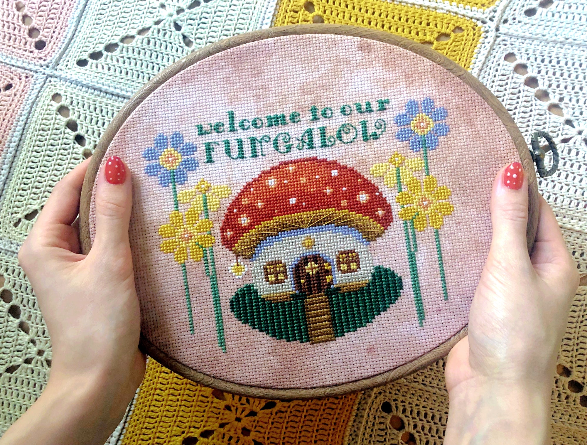 Embroidery hoop held in hand, to show scale. Pattern is of medium size. Backstitches in the toadstool top are clearly visible here. Quote also contains backstitches and fractional stitches. Behind the scene is a crochet blanket in light colors.