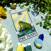 Flatlay of The Friend frog tarot cross stitch pattern. Stitched item, surrounded by decorations. Finished piece is of small to medium size. Colors are yellow, green, blue, grey and black. Frog is cute and small. The Friend is written underneath.