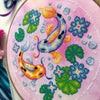 Closeup of Koi Fish cross stitch pattern. Right side is most visible. Finished piece is of medium size. Colors are very bright, and are green, blue, orange, pink and yellow. There are two fish, surrounded by lotus leaves and flowers.