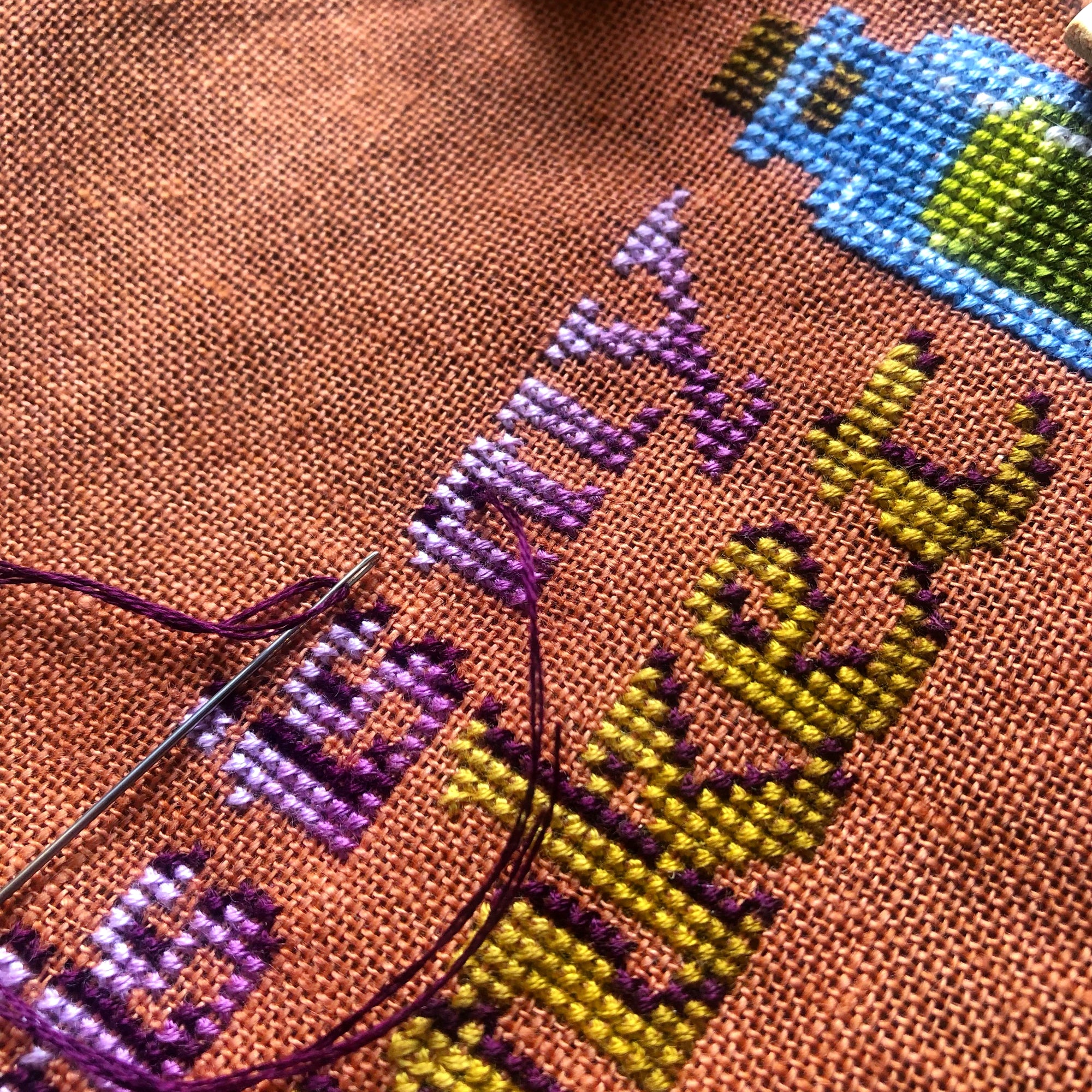 Closeup of the Trinket Bag cross stitch pattern being stitched. Stitches are neat and beautiful, colors really pop. Needle with thread is visible, and is placed on top of the work in progress.